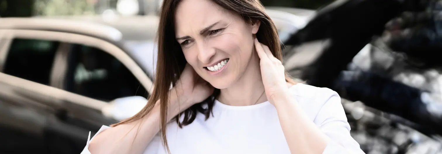 Woman standing outside of her car after.an accident, grimacing in pain as she feels her neck.