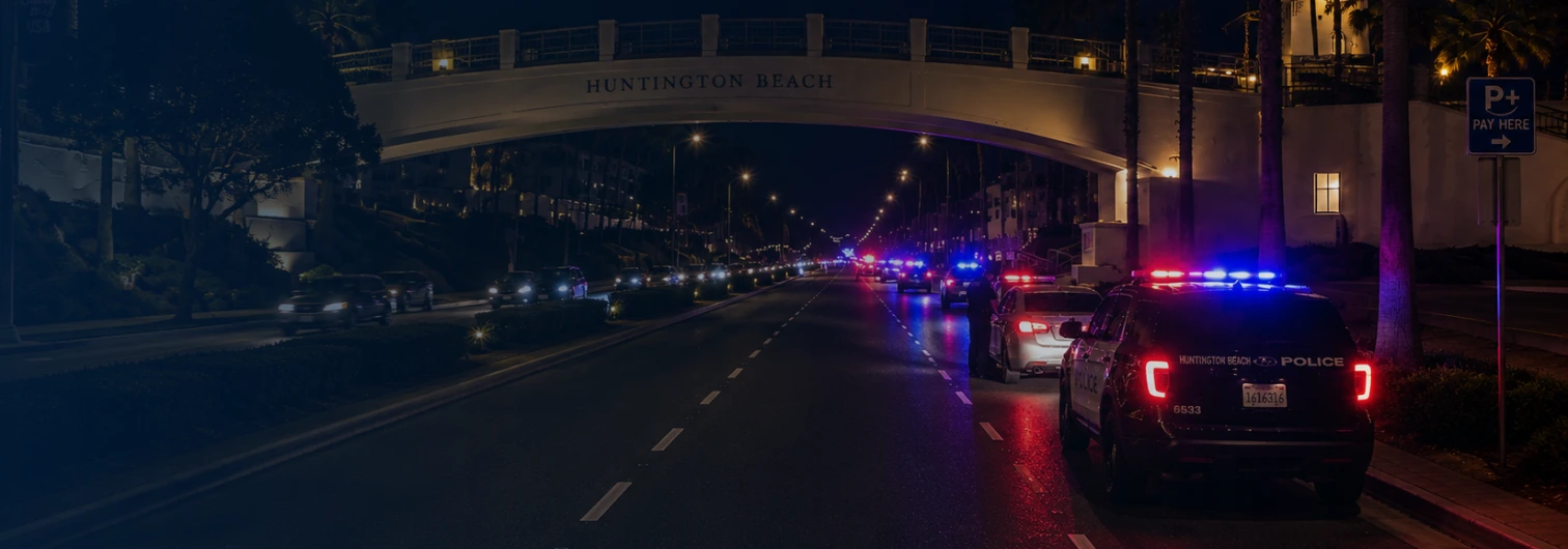Pacific Coast Highway at night near downtown Huntington Beach with red and blue emergency lights visible in the distance