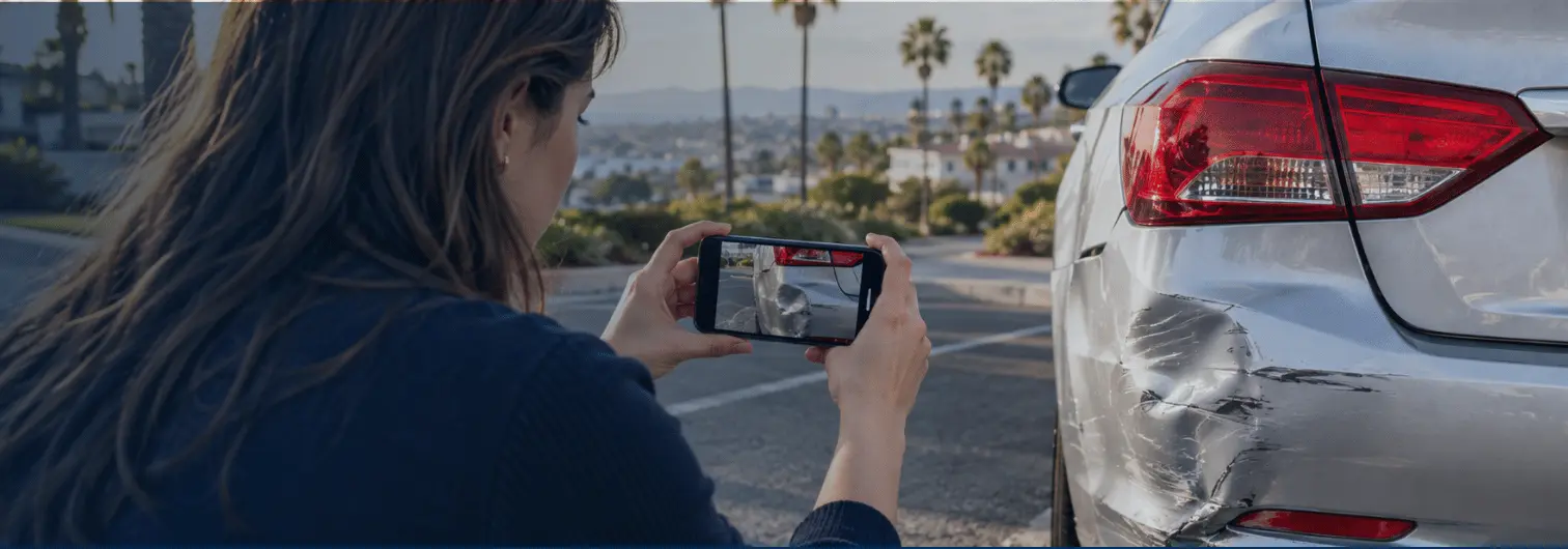 Woman photographing a silver sedan’s damaged rear bumper with her smartphone.